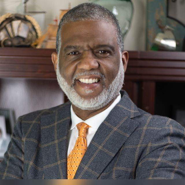Joe Follette smiling, wearing a navy checked suit and orange tie